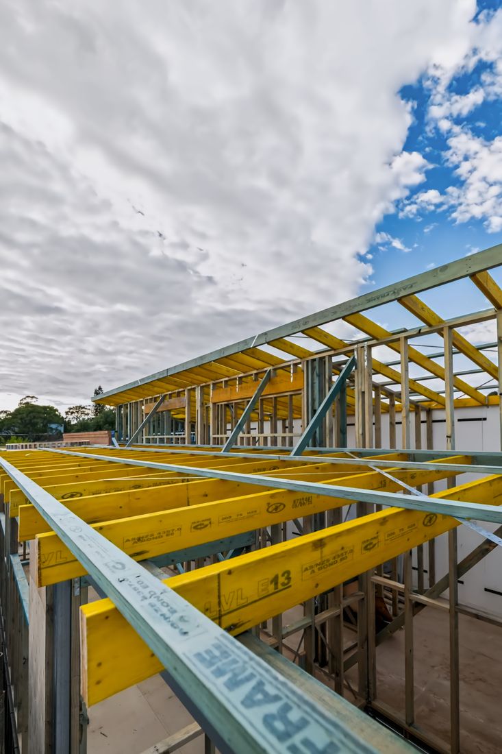 Low-angle view up into a new roof frame of yellow LVL beams braced with galvanised steel, meeting at the ridge against a clouded sky