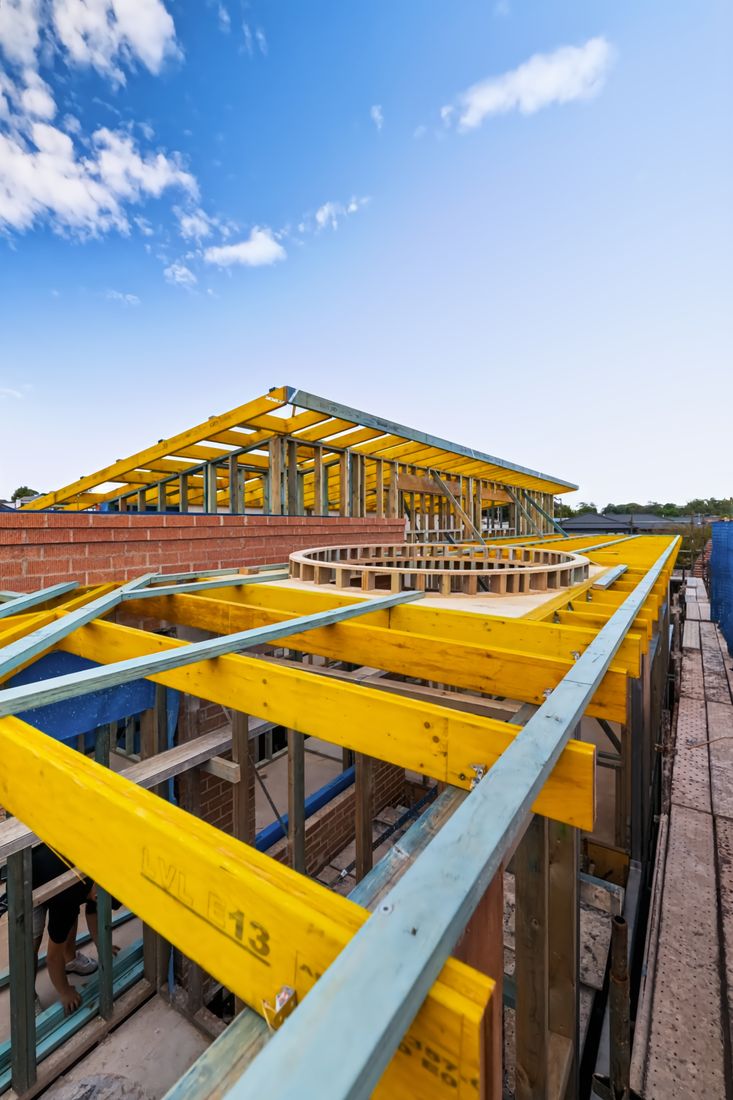 High-angle view across the roof framing, showing yellow LVL beams, a circular opening set in the roof plane, brickwork and scaffold