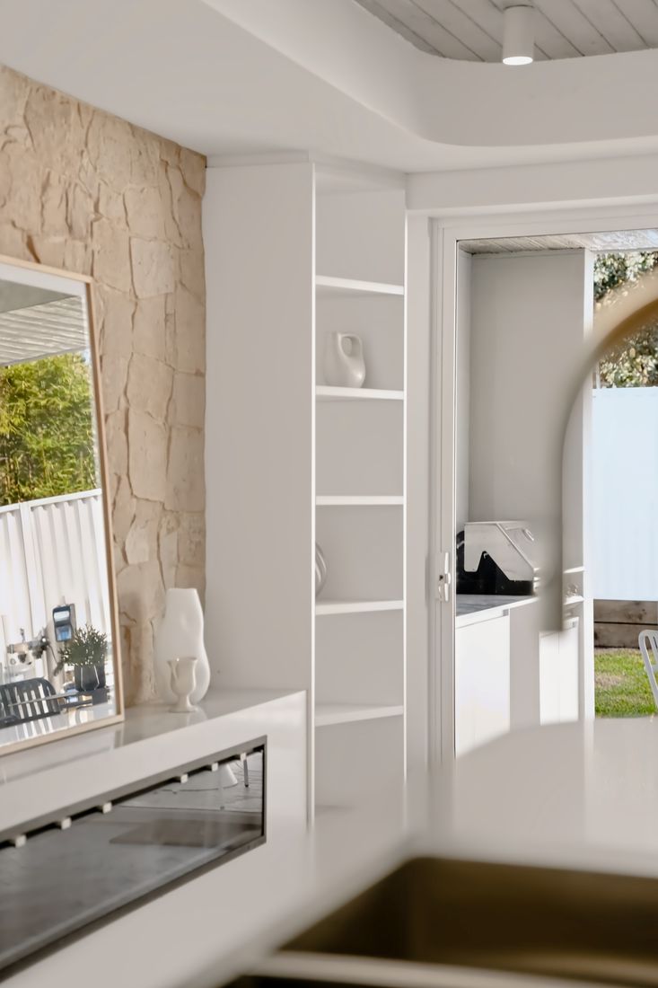 A rough-hewn sandstone feature wall beside a tall white open shelving unit and a large mirror in a light timber frame, with a doorway to the garden