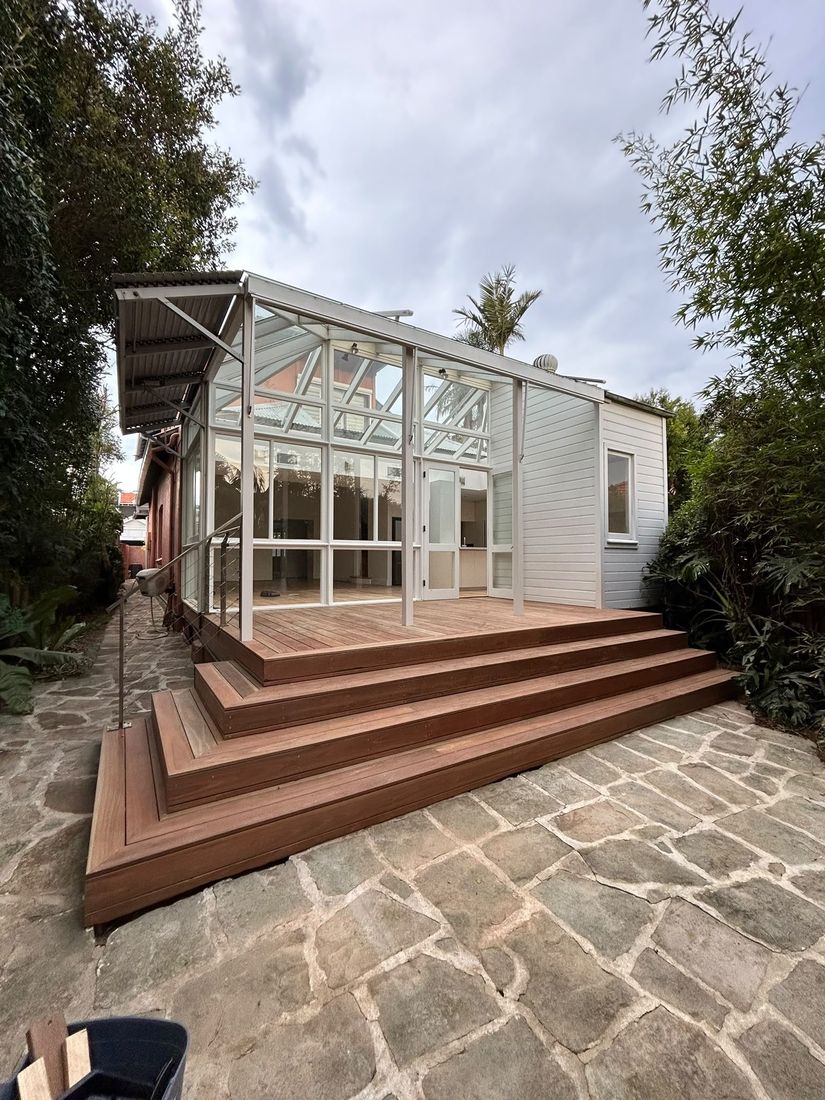 New spotted gum deck and wraparound steps beside a glazed conservatory, over a sandstone-paved courtyard