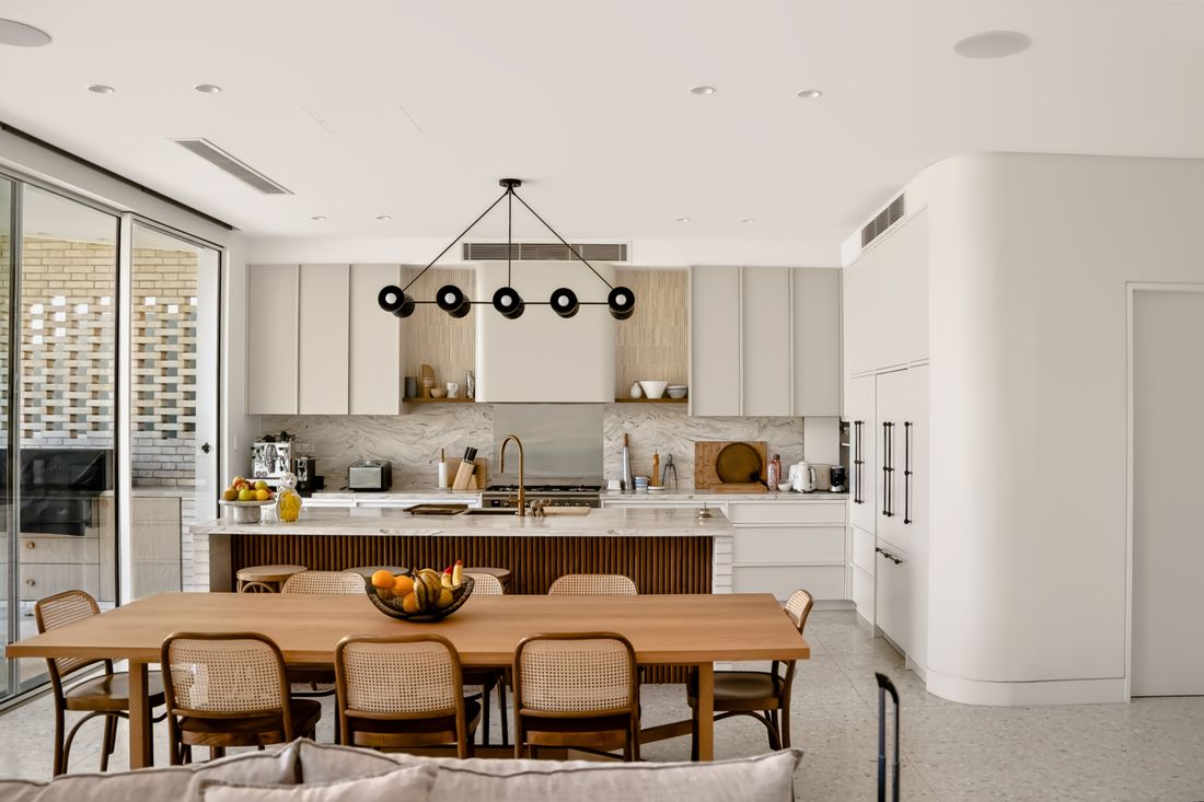Open-plan kitchen and dining area with a marble waterfall island on a dark slatted base, matte white cabinetry and a light oak table with cane chairs