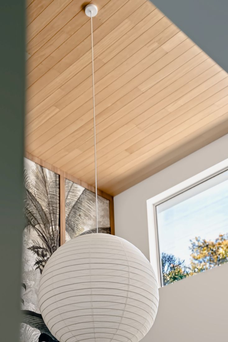 A large white paper lantern hanging from a slatted timber ceiling beside botanical wallpaper and a window