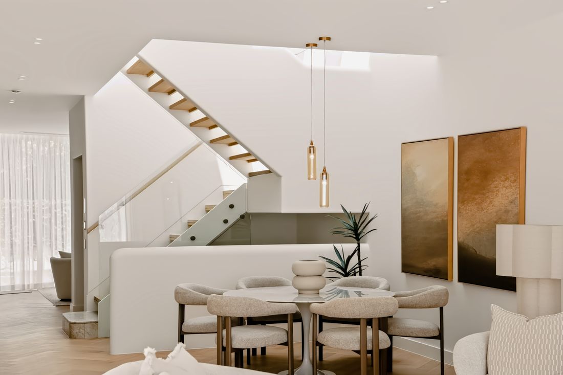 A circular glass dining table with upholstered chairs beneath two brass pendants, with a timber and glass staircase beyond