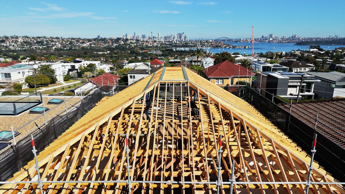 A drone view of the pitched timber roof frame in Dover Heights, with the harbour and city behind
