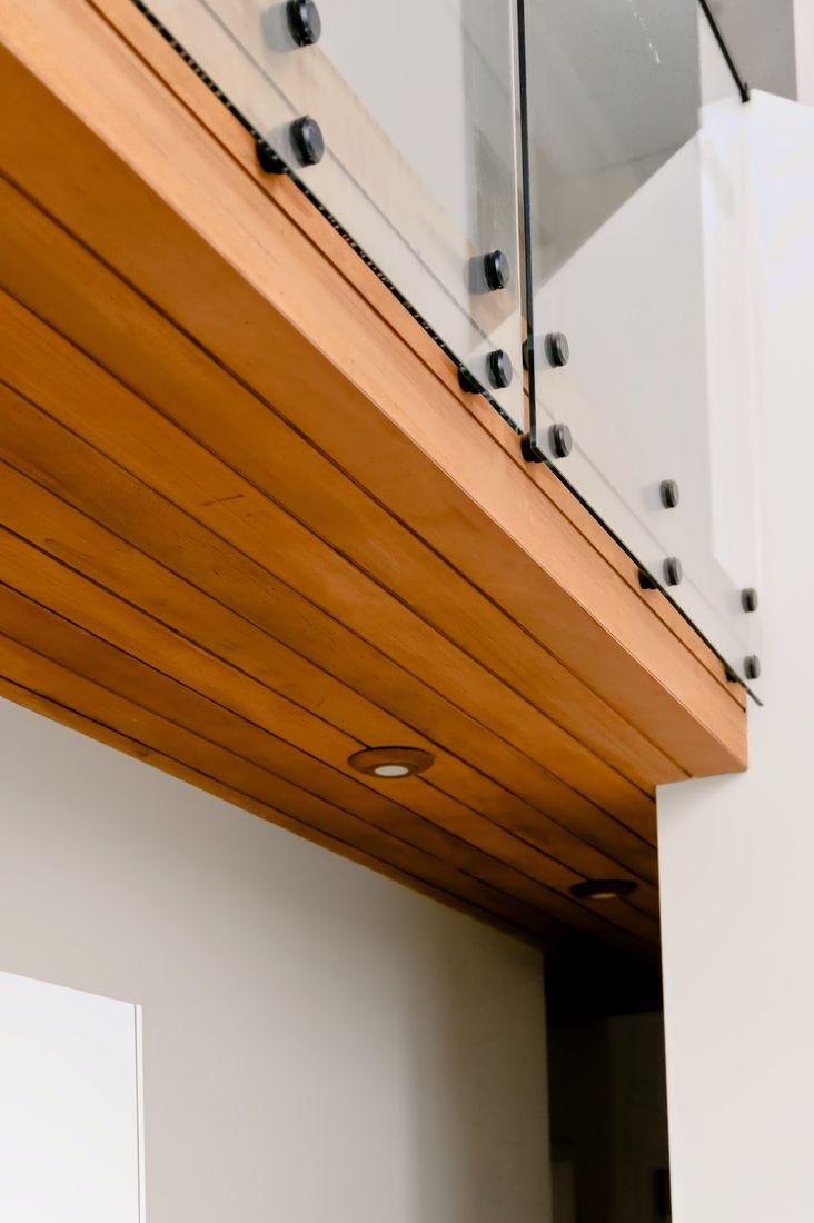 Low-angle view of a loft edge clad in horizontal timber boards, with a glass balustrade fixed by circular black stand-offs and downlights set into the soffit