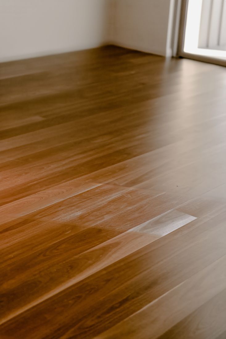 Close view across a polished hardwood floor, its grain and knots catching a broad reflection of daylight, with a pale wall beyond