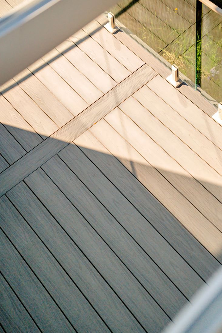 High-angle view across a pale grey wood-grain deck, a framing board dividing the layout, with a glass railing on silver brackets at the edge