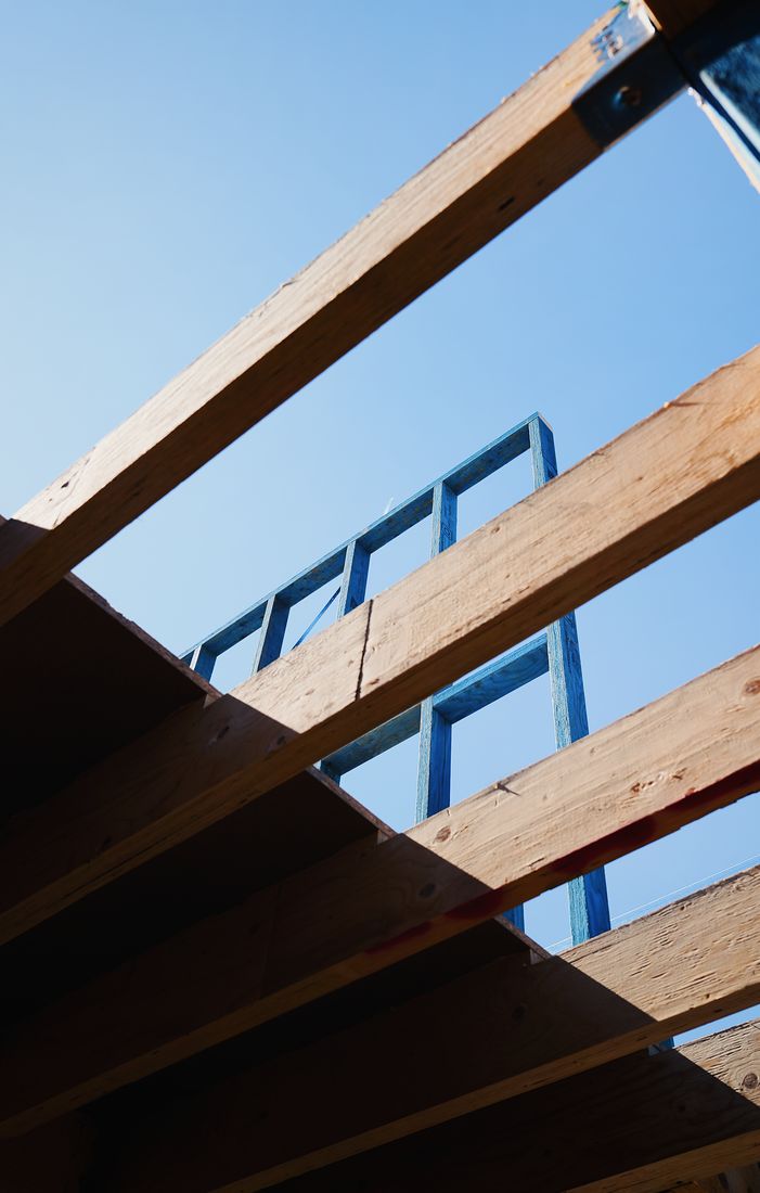 Parallel timber floor joists running diagonally toward a blue-painted wall frame standing against a clear sky, their shadows falling across the floor