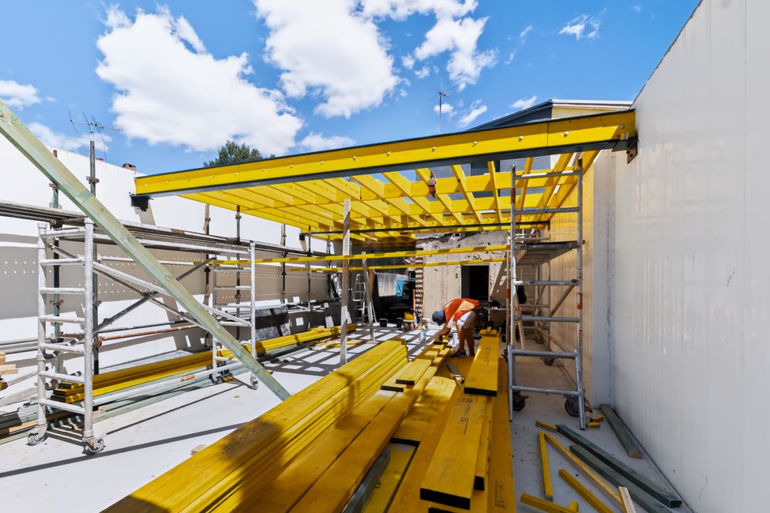 Yellow timber beams stacked on a concrete slab beneath a framed ceiling, with a worker on site