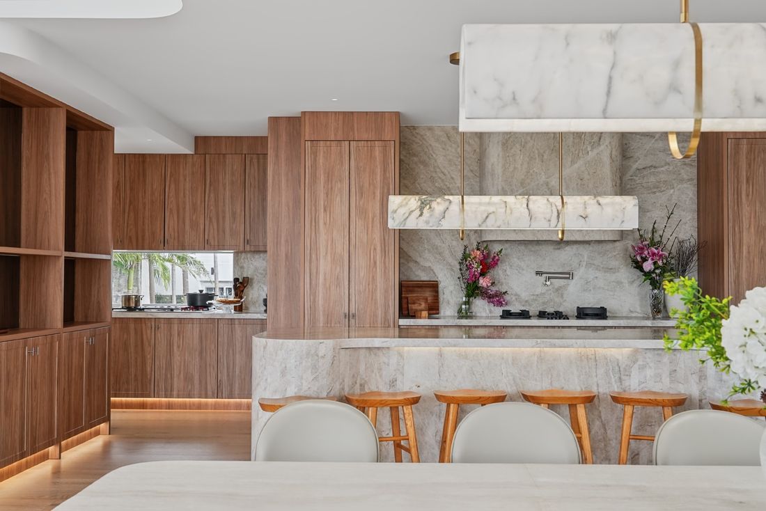 An oak kitchen with a long stone island and timber bar stools, a stone splashback and floating stone shelf, and a marble pendant light above